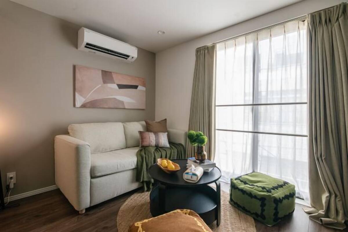 A living room with a sofa, a round coffee table, a small ottoman, a potted plant, and a window with curtains.