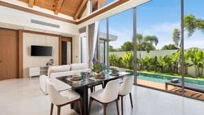 A modern living room with a dining area, large windows, and a view of a swimming pool. - The Ozon Bangtao 3-bedrooms Villas with pool by My Home Phuket (Phuket)