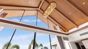 A modern interior with a high ceiling, wooden beams, and large windows offering a view of palm trees. - The Ozon Bangtao 3-bedrooms Villas with pool by My Home Phuket (Phuket)