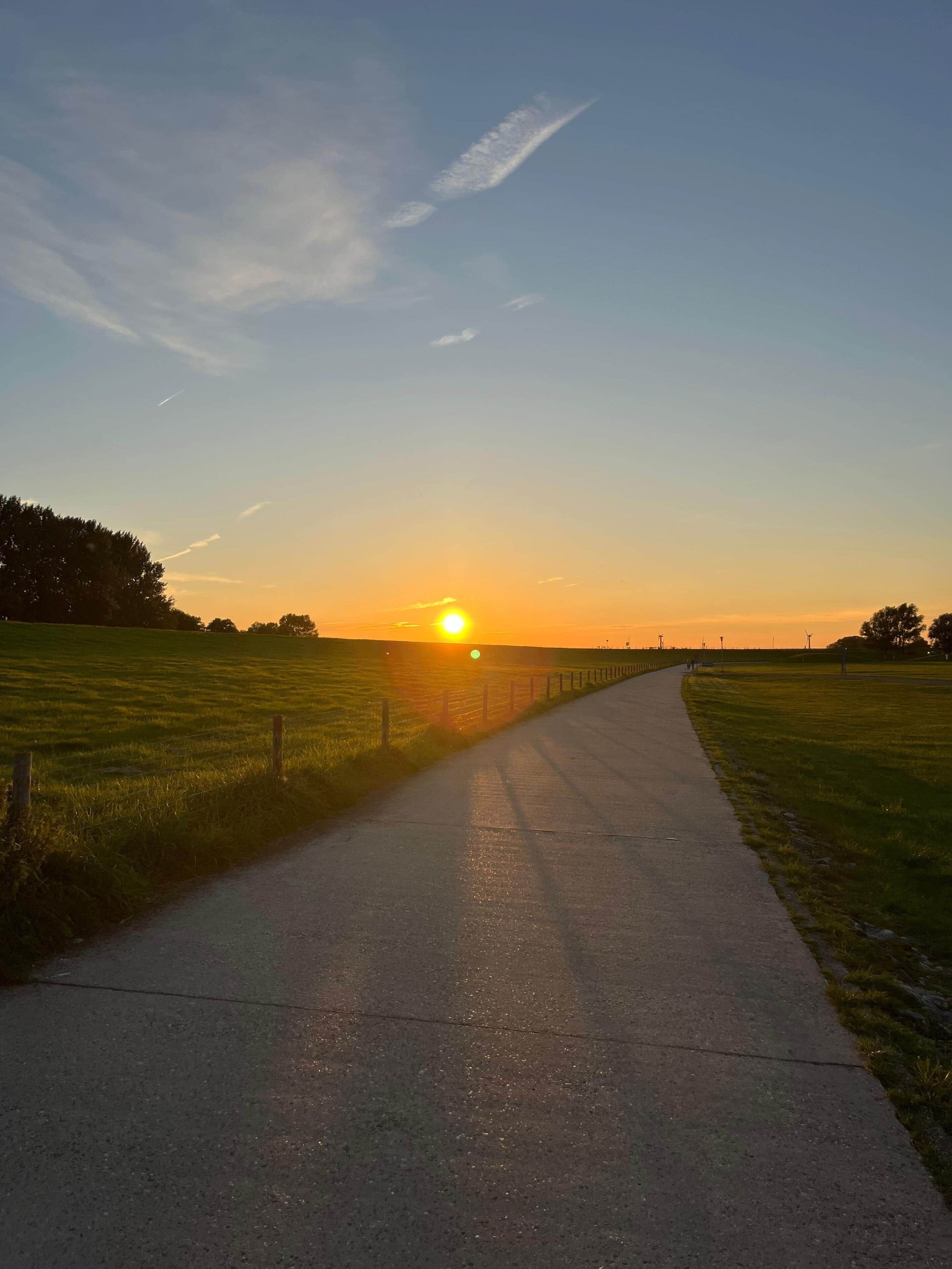 Een verharde weg die richting een zonsondergang leidt, met een hek aan één kant en open velden aan de andere kant.
