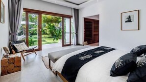 A modern bedroom with a large bed, a seating area with a chair, and a view of a lush garden through the sliding glass doors.