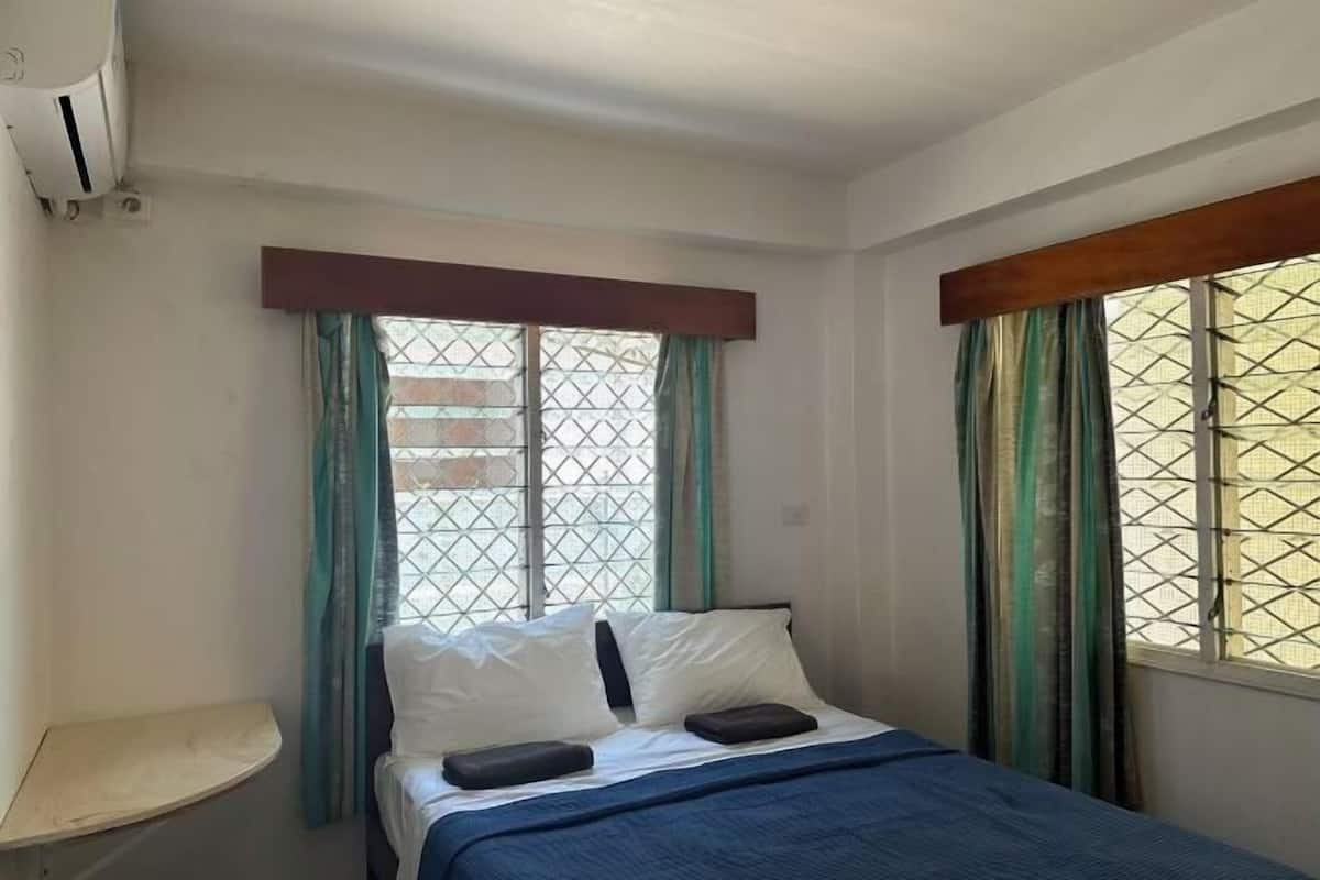 A single bed with a blue bedspread, two pillows, and a wooden headboard. There is a small bedside table and a window with curtains.