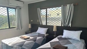 A hotel room with two beds, each with a patterned bedspread, a nightstand with a lamp, and a window with curtains. - Ac airport apartment (Nadi)
