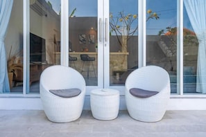 Two wicker chairs with cushions, a small round table, and a glass door leading to an outdoor area with a dining table and chairs. - Villa Casa Antonio (Bali)