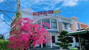 Ein Hotel namens „SAIGON HA TIEN HOTEL“ mit blauem Himmel, einem blühenden Baum im Vordergrund und einer gepflegten Außenansicht.