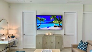 A living room with a television displaying a tropical beach scene, a desk with a lamp, a sofa with patterned cushions, and a cabinet with candles.