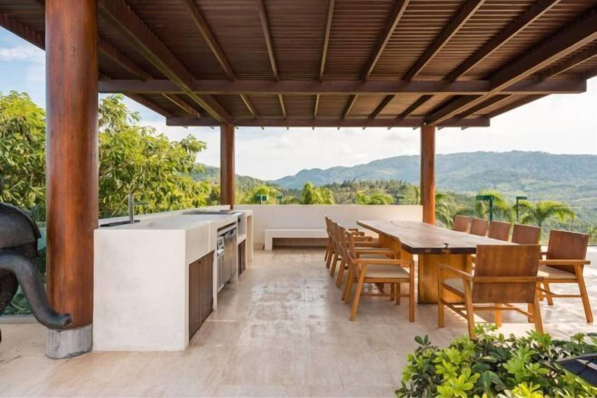 An outdoor patio with a wooden table and chairs, a grill, and a view of the mountains.