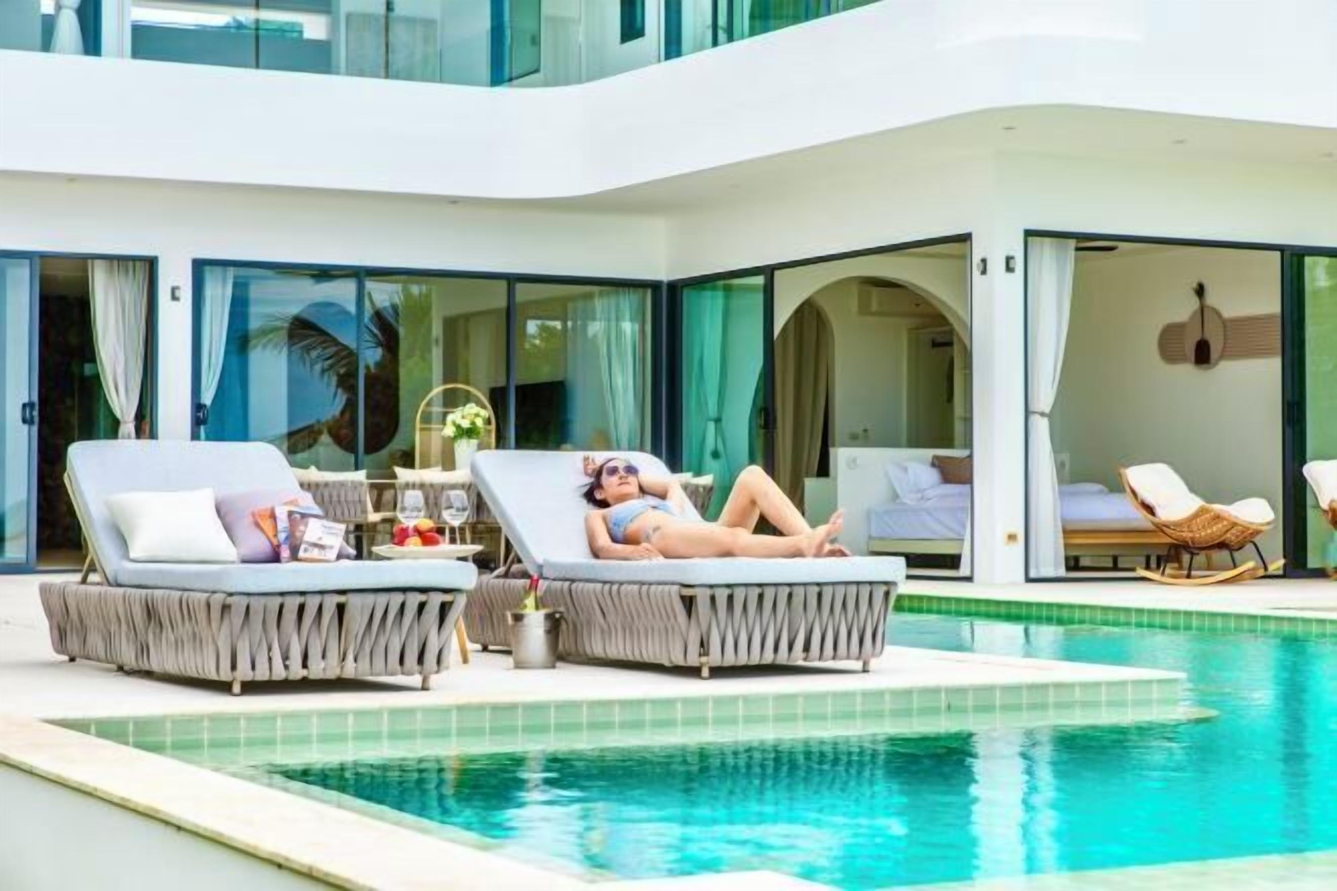 Un área moderna junto a la piscina con dos sillas de descanso y una mesita, vista despejada de la piscina y un vistazo al interior a través de amplias puertas de vidrio.