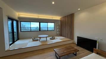 Quarto moderno com uma janela ampla, televisão de tela plana, um banco de madeira e uma pequena mesa redonda.