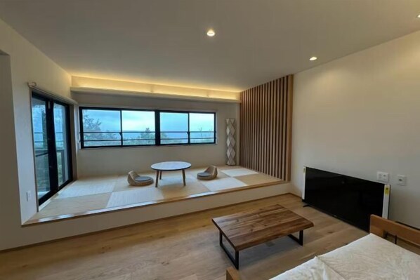 A modern room with a large window, a flat-screen TV, a wooden bench, and a small round table. - LAGUNA OKINAWA Beachfront YAKA (Kin)