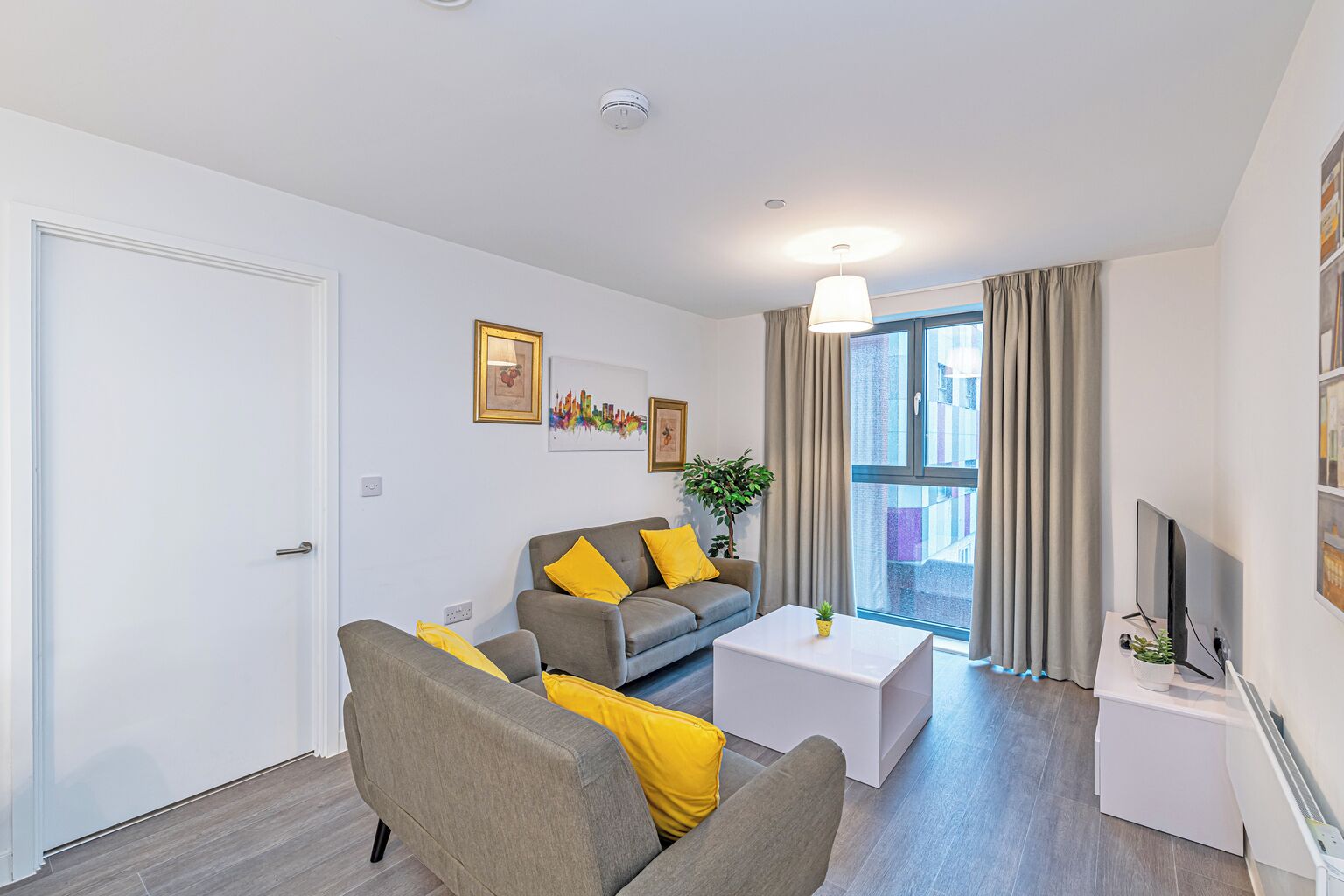 A modern living room with a grey sofa, yellow cushions, a white coffee table, and a TV on a white stand.