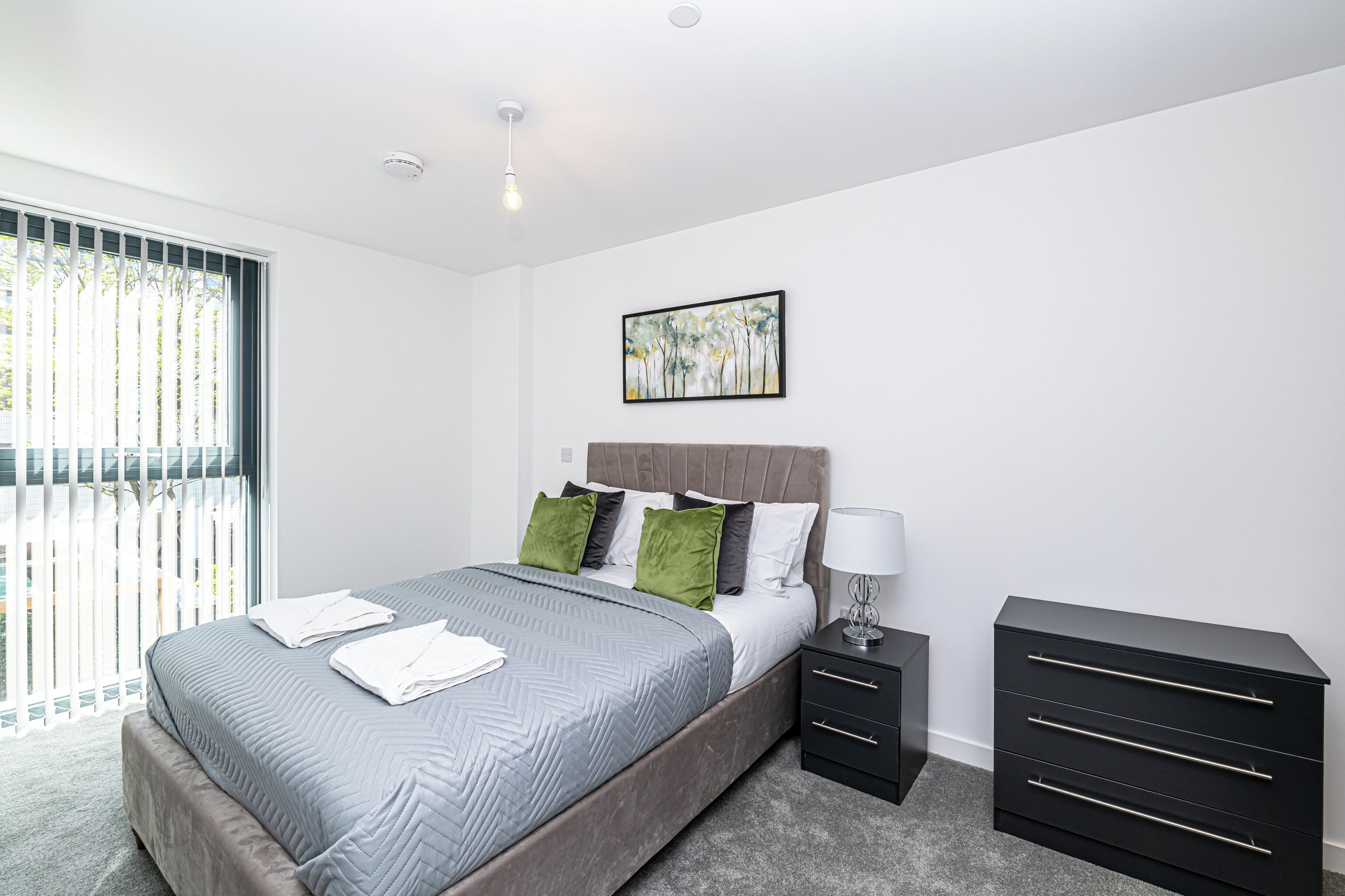 A modern bedroom with a large bed, a nightstand, a dresser, and a ceiling light.