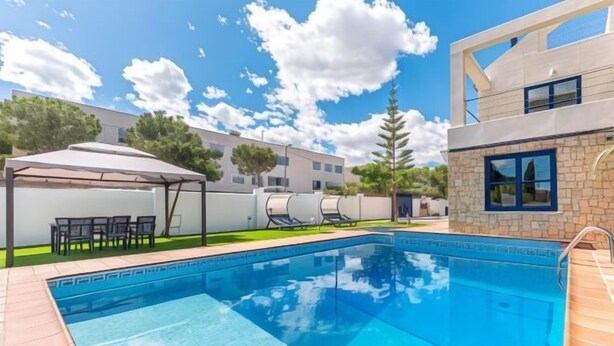 A swimming pool with a tiled deck, a covered seating area, and a modern building in the background. - Villa Lucentum (Alicante)