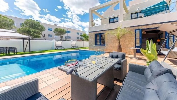 A pool area with a table and chairs, a lounge chair, and a building with a balcony. - Villa Lucentum (Alicante)