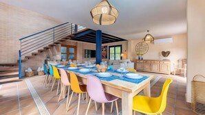 A spacious dining area with a long wooden table set for six, colorful chairs, and a staircase leading to an upper level. - Villa Lucentum (Alicante)