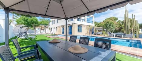 An outdoor patio with a table and chairs, a pool in the background, and a building with a poolside area.