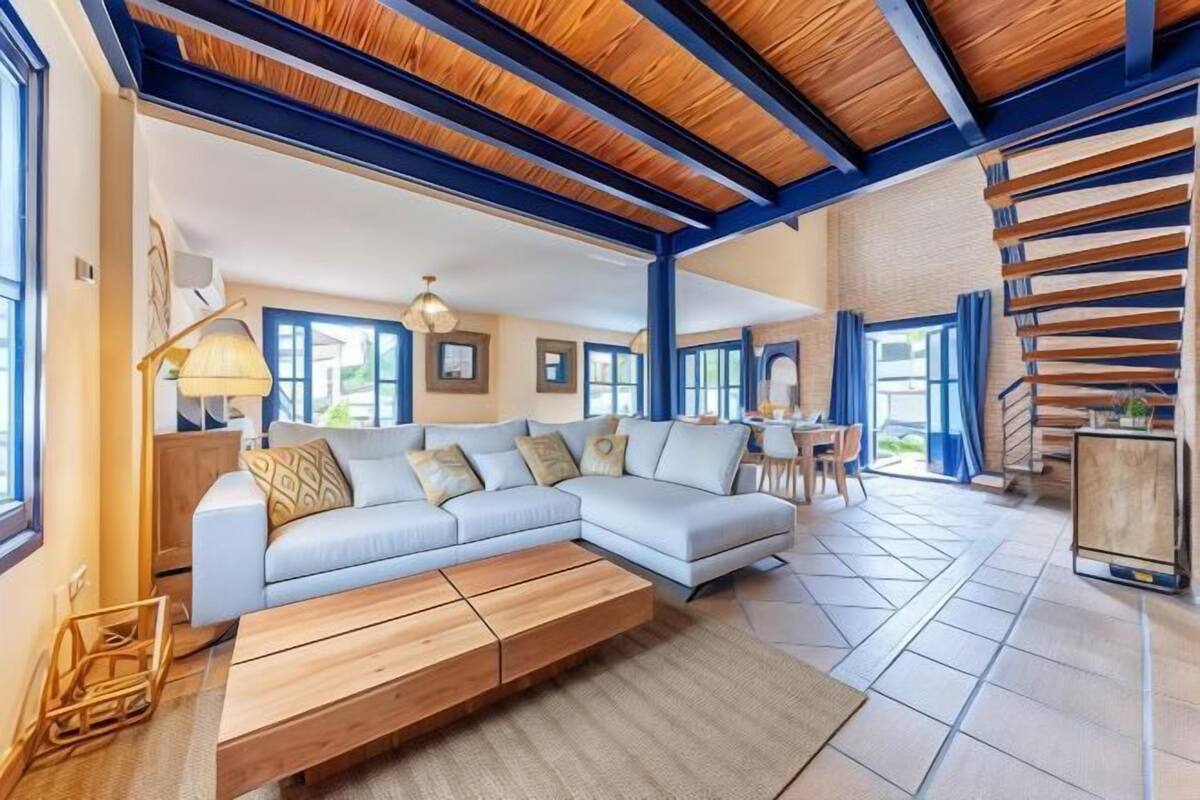 A spacious living room with a light-colored sectional sofa, a wooden coffee table, and a dining area with a small table and chairs.