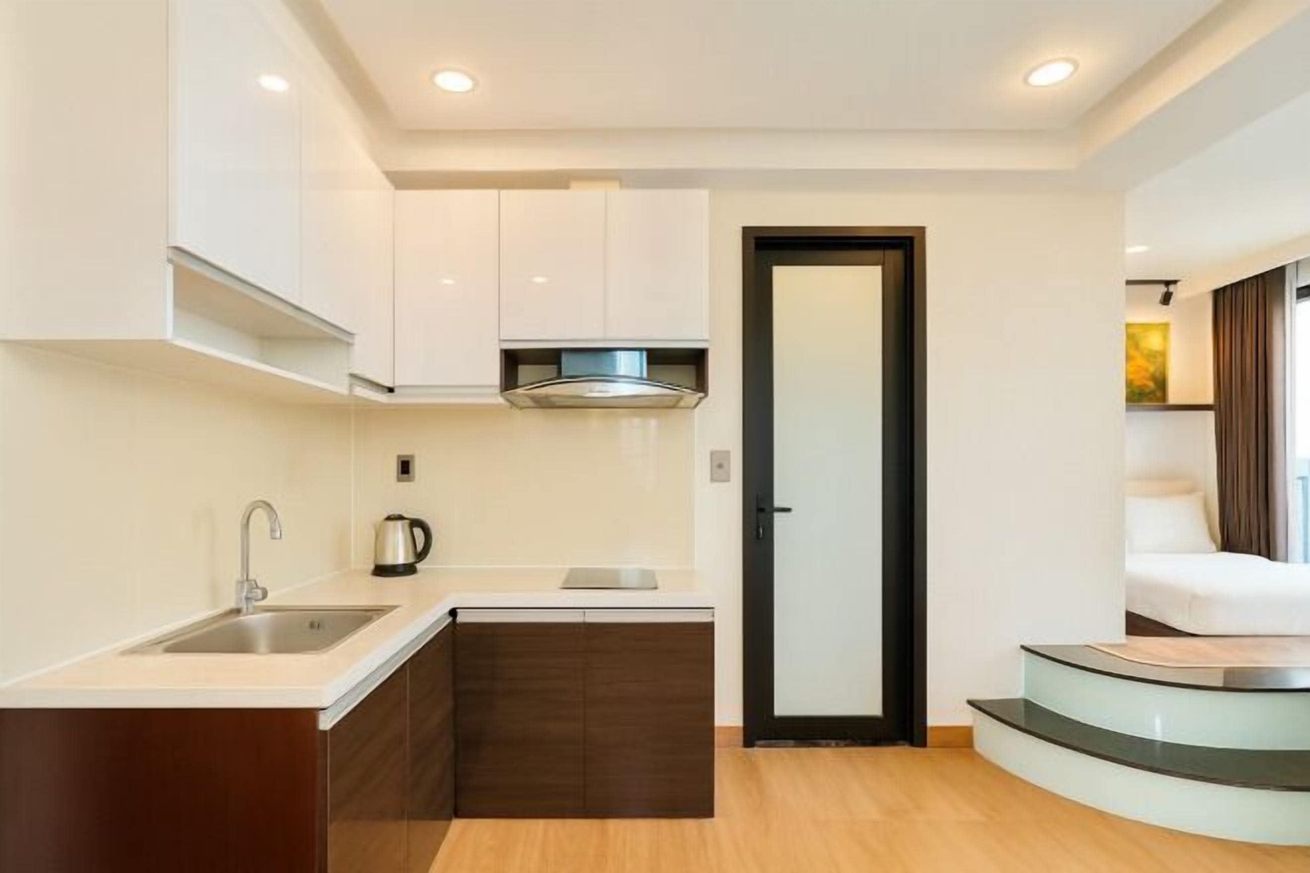 A compact kitchen with a sink, stove, and cabinets, leading to a bedroom with a glass staircase.