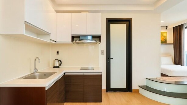 Une cuisine compacte équipée d’un évier, d’une cuisinière et de placards, qui donne sur une chambre à coucher avec un escalier en verre.
