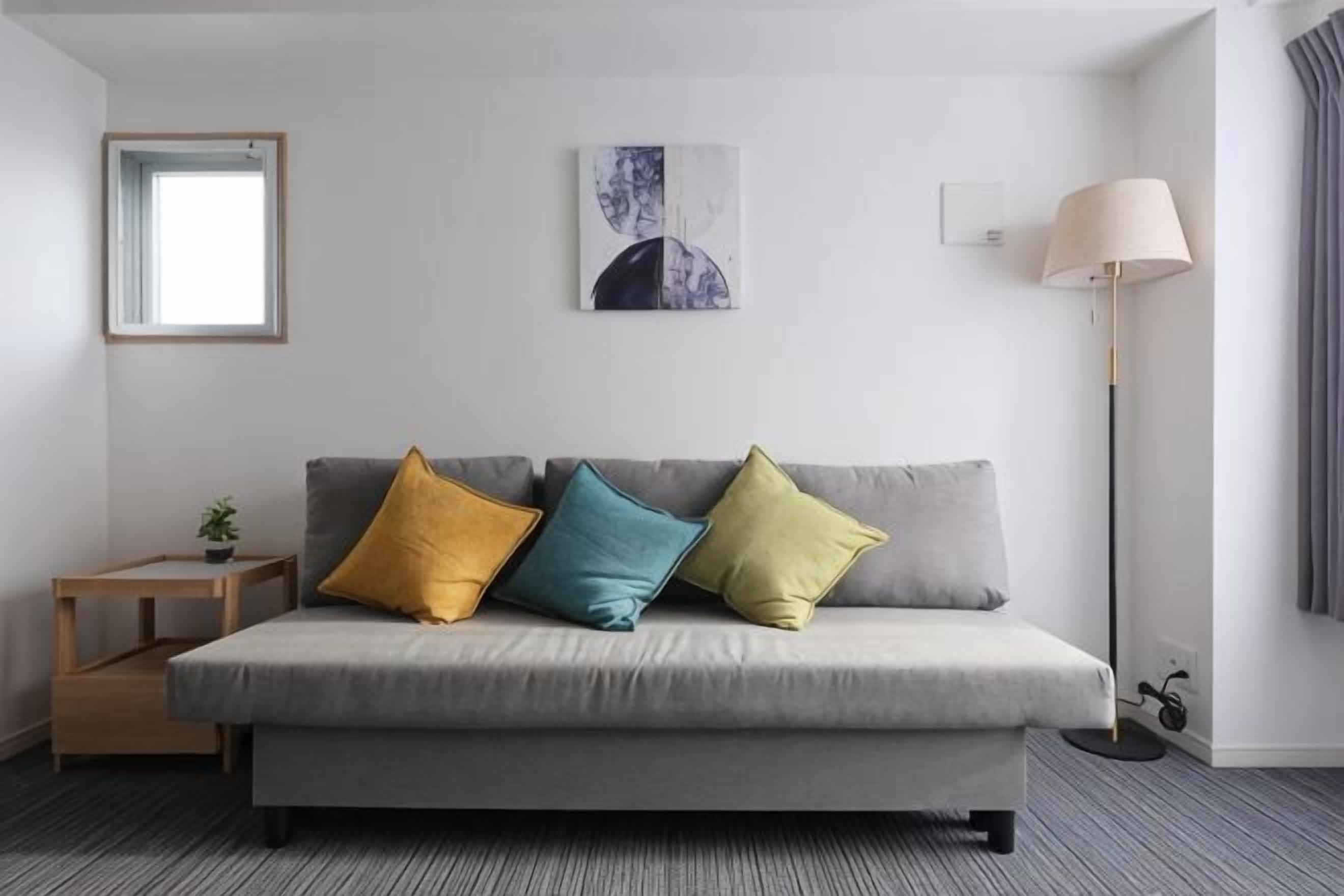 A neutral-colored sofa with three cushions, a small wooden side table with a plant, a floor lamp, and a framed picture on the wall.
