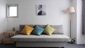 A neutral-colored sofa with three cushions, a small wooden side table with a plant, a floor lamp, and a framed picture on the wall.