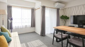 Une chambre d’hôtel avec un canapé, une table à manger avec des chaises, une télévision et une grande fenêtre avec des rideaux.