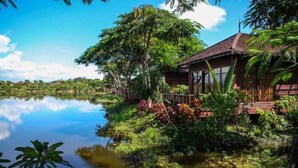 A wooden house with a thatched roof, surrounded by lush greenery and a calm lake.