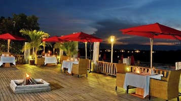 An outdoor dining area with tables and chairs, red umbrellas, and a fire pit.