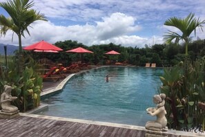 Ein Swimmingpool mit Liegestühlen und Sonnenschirmen, umgeben von tropischer Bepflanzung.