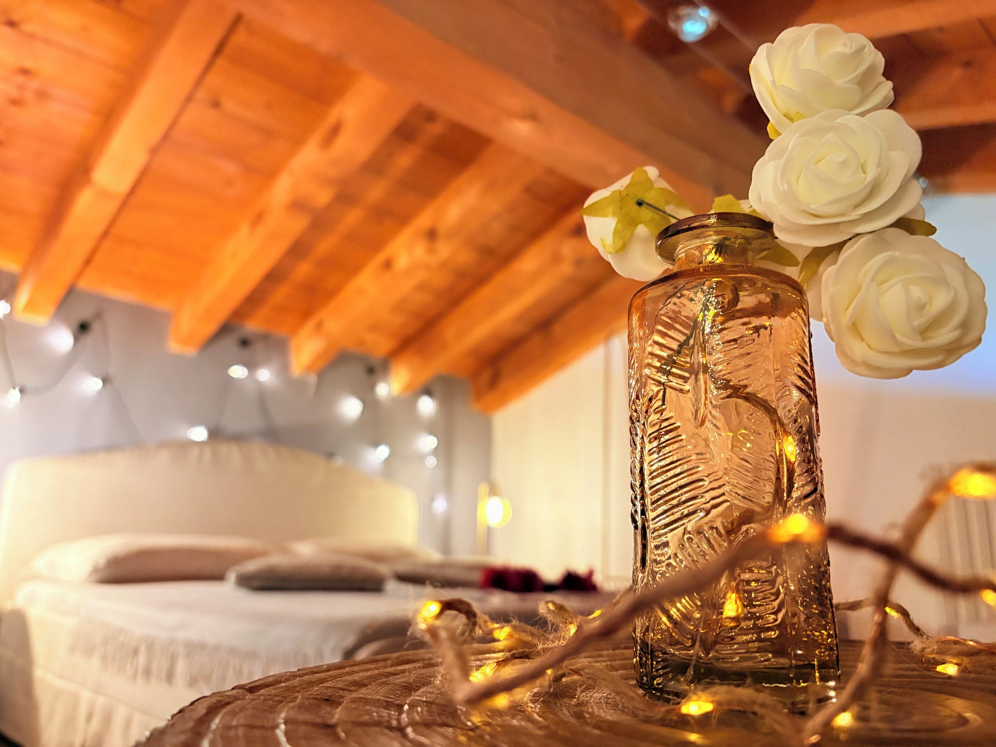 Ein gemütliches Schlafzimmer mit Holzbalkendecke, einem Bett mit weißer Bettwäsche und einer Glasvase mit weißen Blumen auf einem runden Tisch.