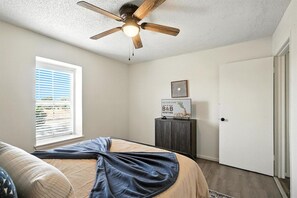 A bedroom with a bed, a ceiling fan, a window with blinds, a door, and a small cabinet with a sign.