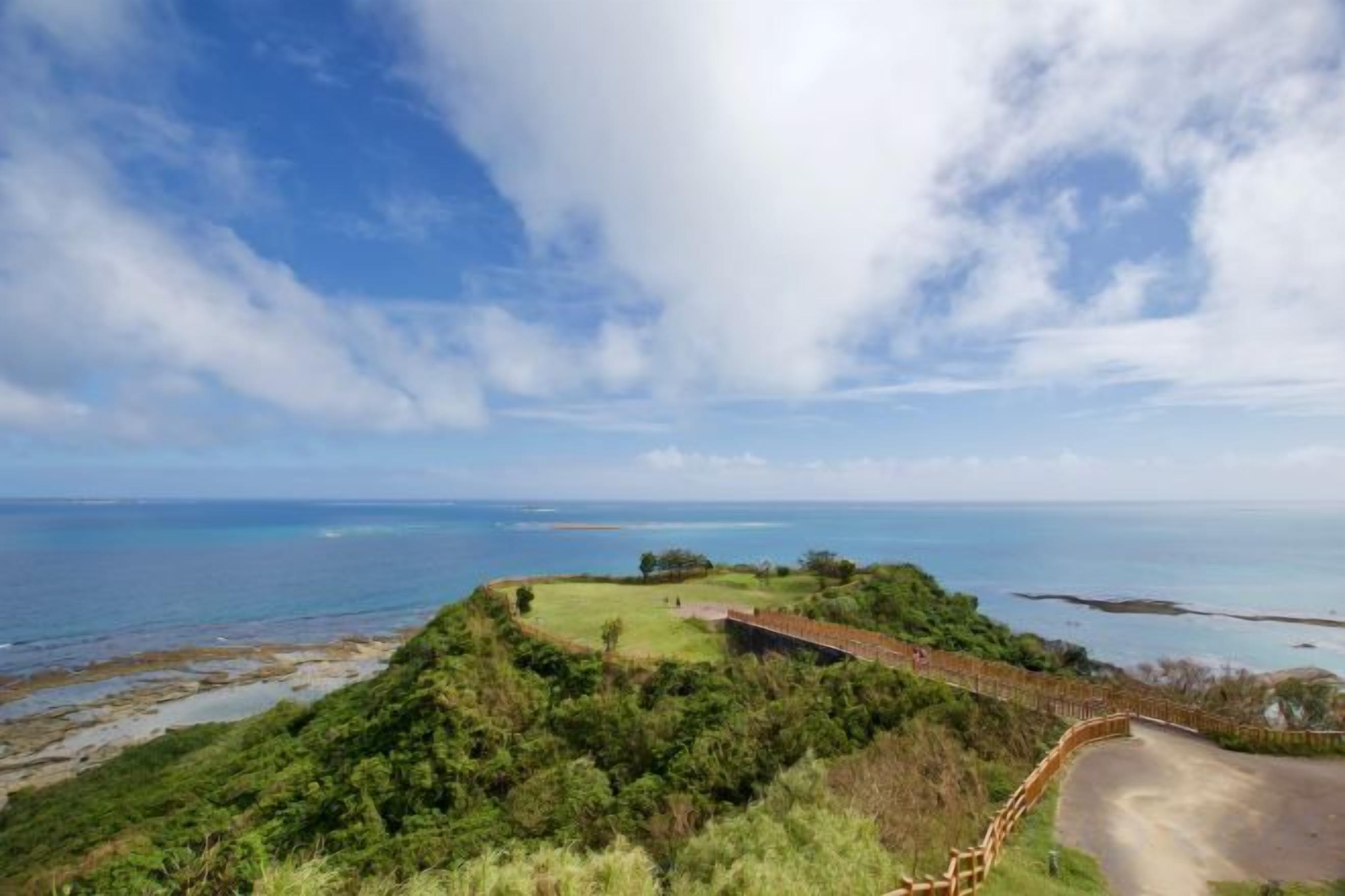 A coastal landscape with a paved path, lush greenery, and a view of the ocean.