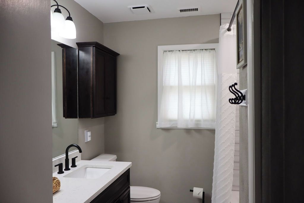A bathroom with a white sink, dark cabinets, a toilet, and a window with sheer curtains.