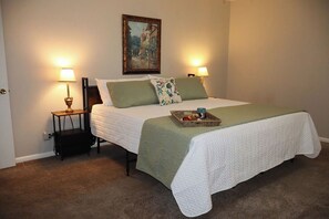 A neatly made bed with a green and white bedspread, two bedside lamps, a small table with a lamp, and a painting on the wall.