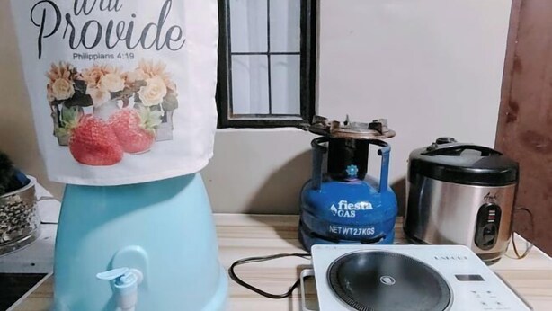 Eine Küchenarbeitsplatte mit Wasserspender in Blau, einer blauen Gasflasche, einem Edelstahl-Reiskocher und einem weißen elektrischen Reiskocher.