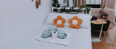 Cama bem arrumada com dois travesseiros em formato de flor laranja, um cobertor branco e um travesseiro xadrez.