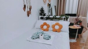 A neatly made bed with white bedding and two orange flower-shaped pillows. There is a small table with a plant and a vase of dried flowers. - Home in Pagadian City near airport (Pagadian)