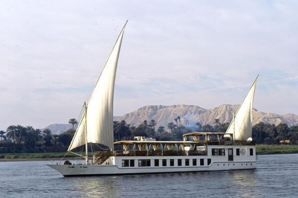 Un barco con dos velas, cubierta protegida y montañas al fondo.