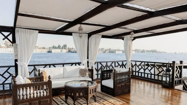 A covered deck with a wooden floor, white curtains, and a view of the water. - Dahabeya Farouz El Nil 3 (Al Bairat)