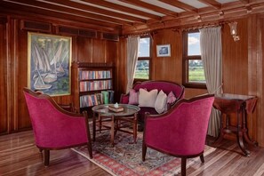 A room with wooden paneling, a bookshelf, a red armchair, a round table, and a view of the outdoors. - Dahabeya Farouz El Nil 3 (Al Bairat)