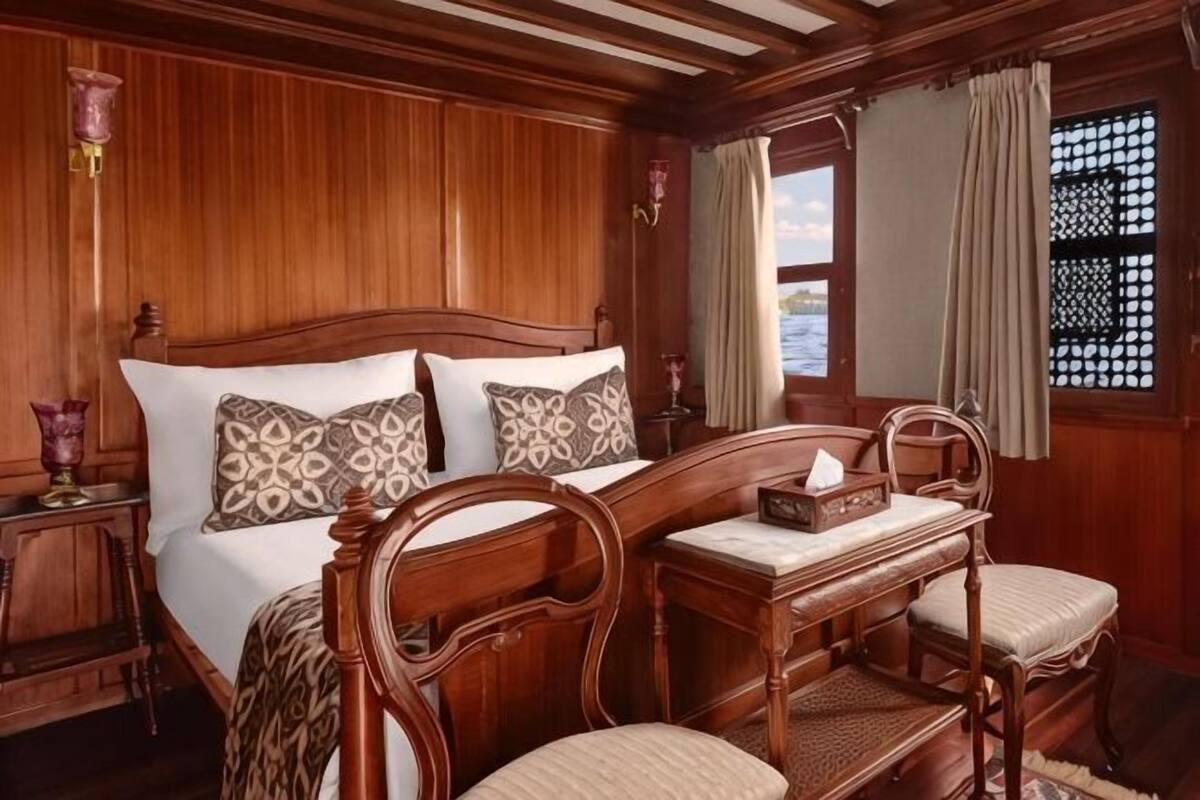 Quarto com painéis de madeira, uma cama, duas cadeiras, uma mesinha de cabeceira e uma janela com cortinas.
