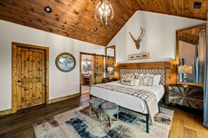 A bedroom with a wooden ceiling, a bed with a tufted headboard, a deer head mounted on the wall, and a rustic wooden door. - Smoky Mtn High: mtn view • pool table • Golden Tee (Gatlinburg)