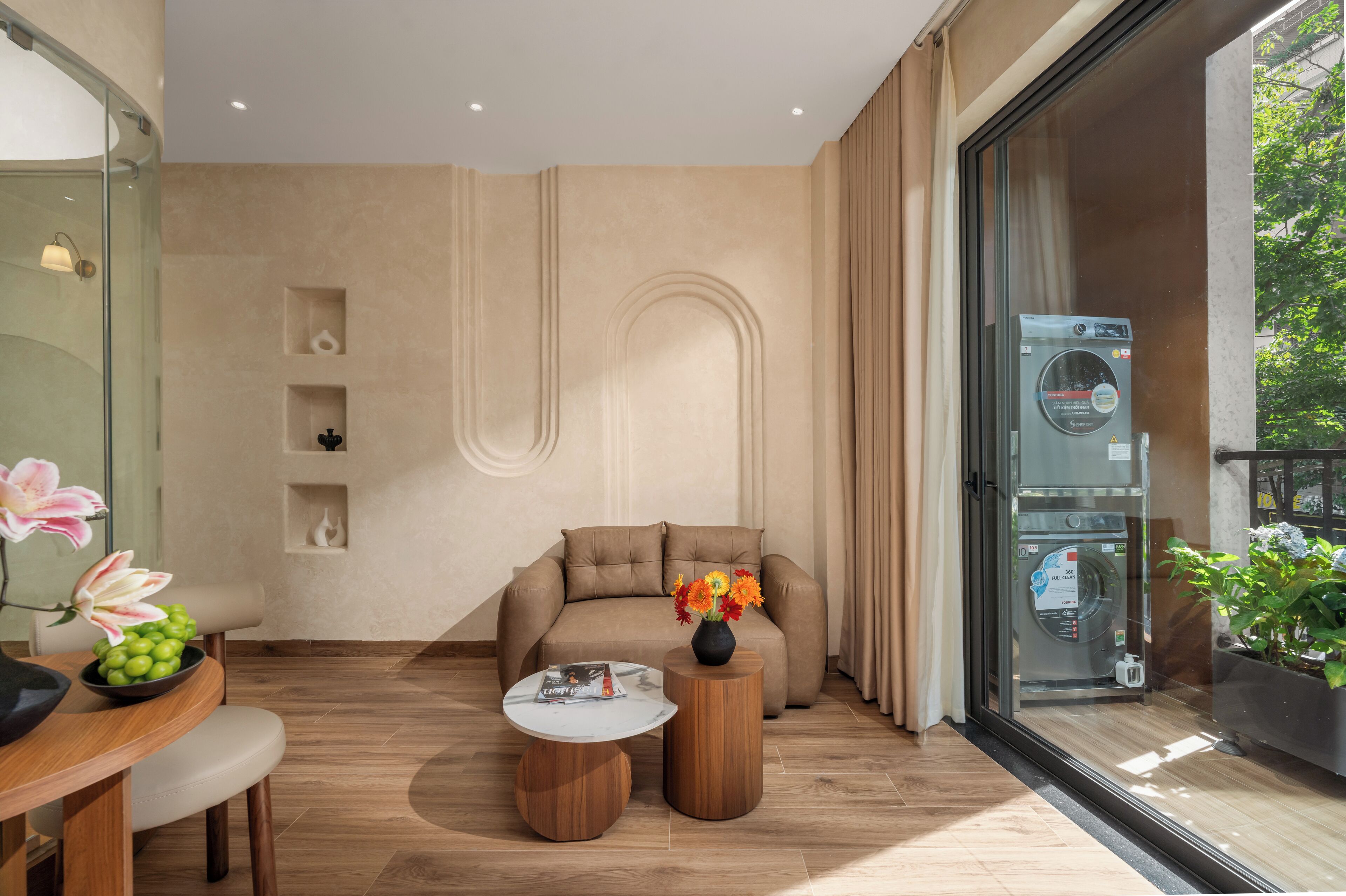 A modern living room with a sofa, a coffee table, and a small table with a vase of flowers. There is a washing machine visible through a glass door.