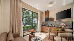Un salon moderne comprenant un canapé, une table basse et un coin repas avec des chaises. On y trouve également une cuisine équipée avec un téléviseur et une grande fenêtre donnant sur un espace verdoyant.