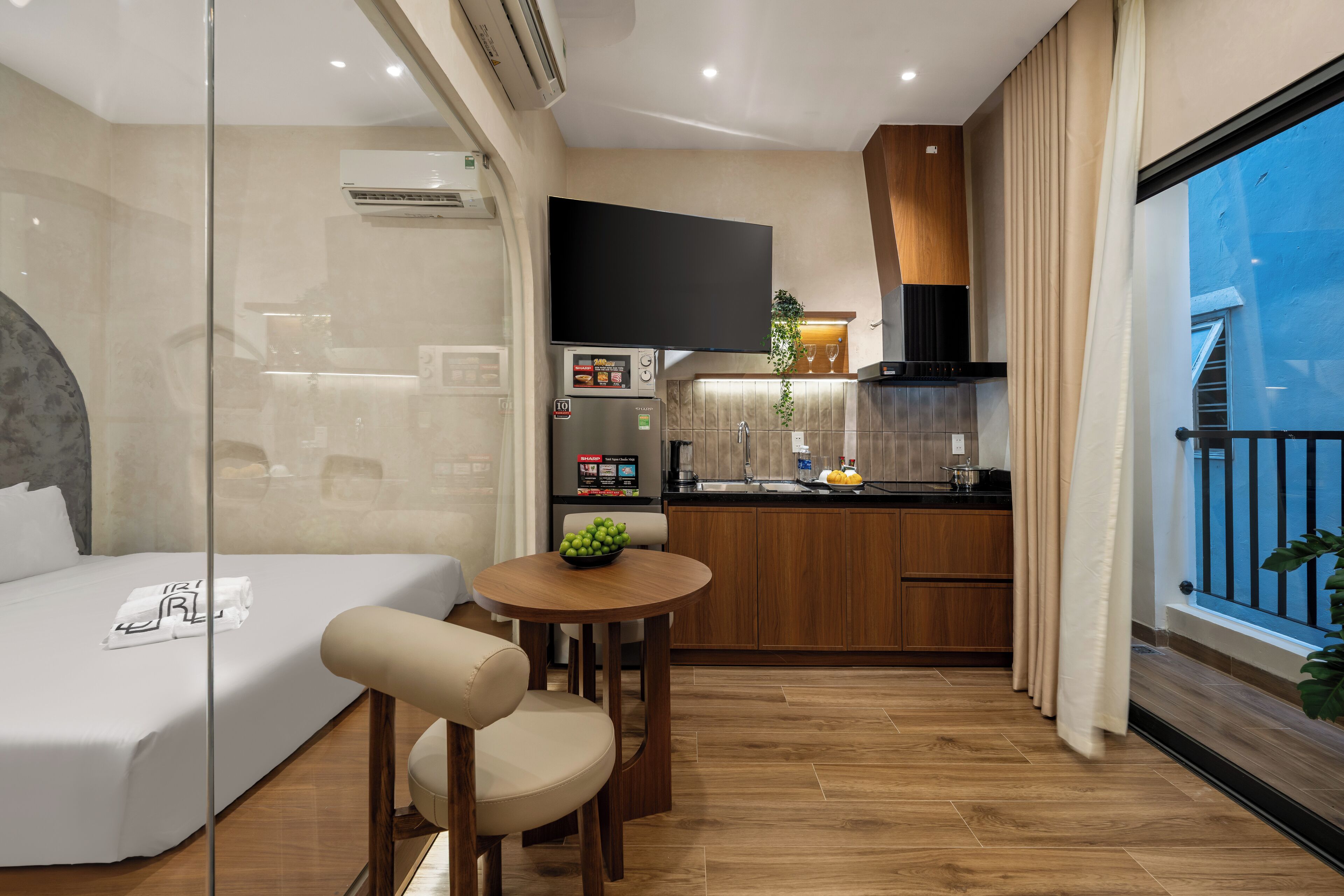 Une chambre d’hôtel avec un lit, une petite table à manger avec un saladier de fruits, un coin cuisine équipé d’un four à micro-ondes et une grande fenêtre avec des rideaux.