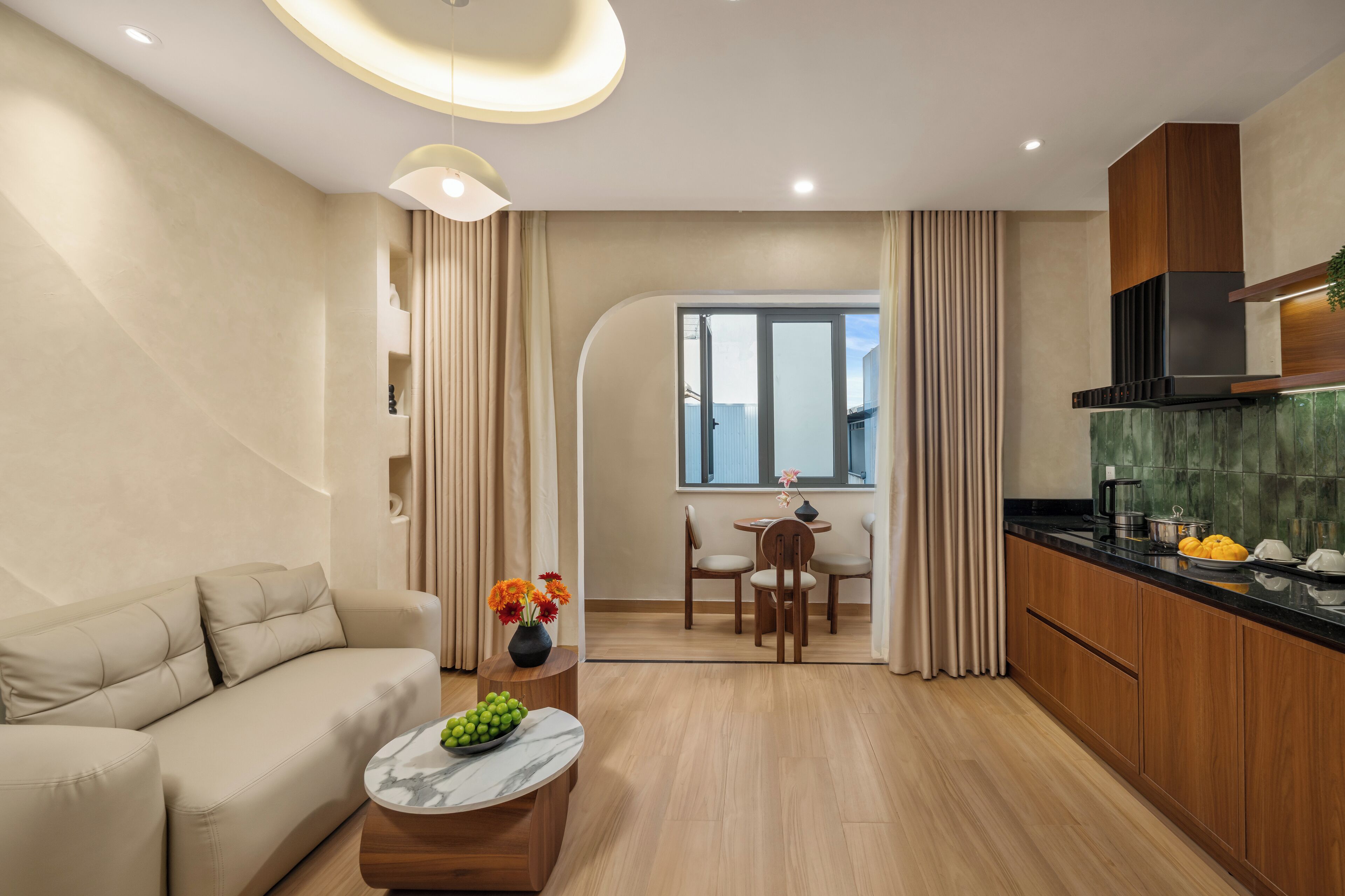 A modern living room with a sofa, a coffee table with fruit, a dining area with chairs, and a kitchen with wooden cabinets and a black range hood.