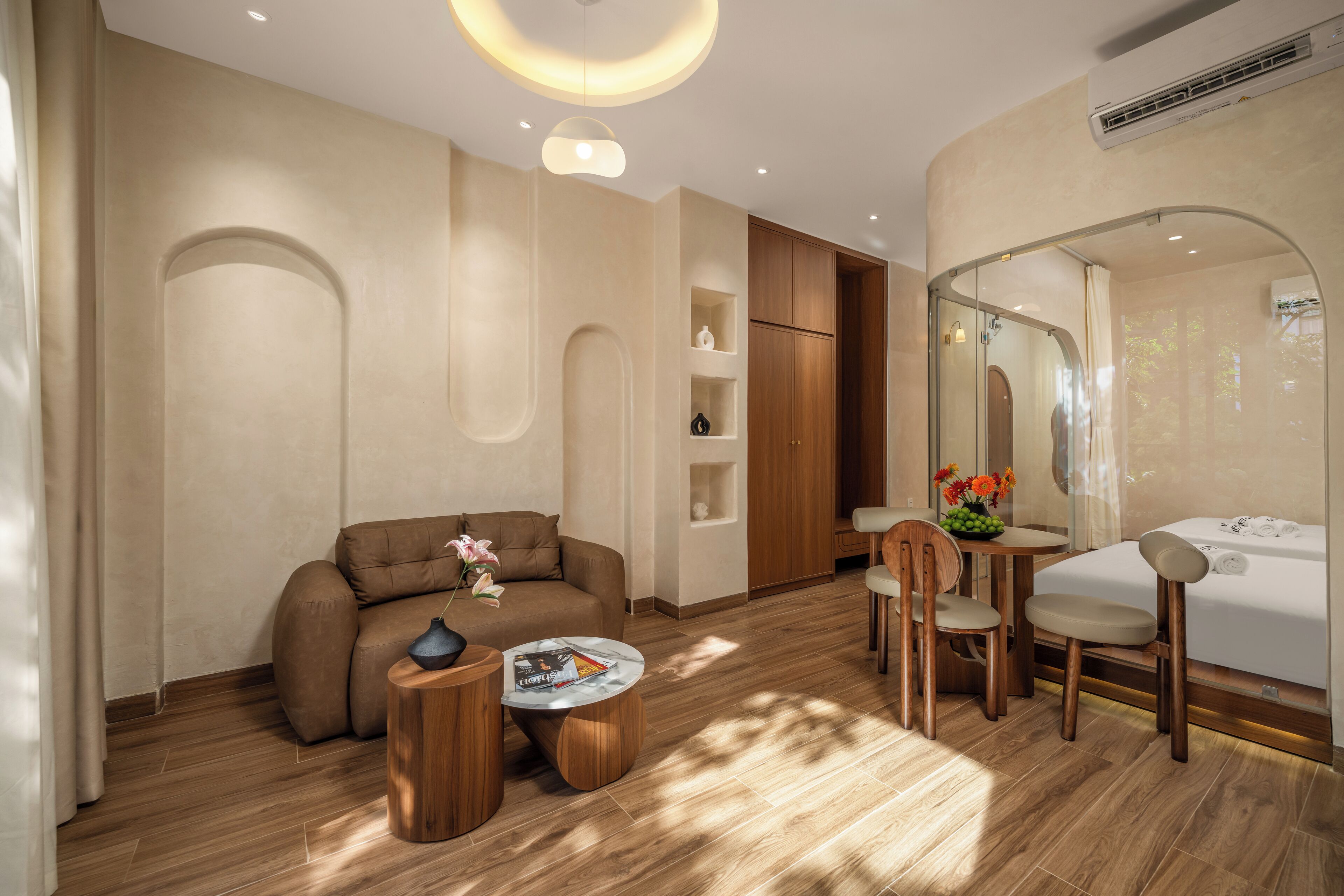 A modern hotel room with a sofa, a small round table, a wooden chair, a glass-enclosed shower, and a wooden cabinet.