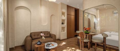 A modern hotel room with a sofa, a small round table, a wooden chair, a glass-enclosed shower, and a wooden cabinet.