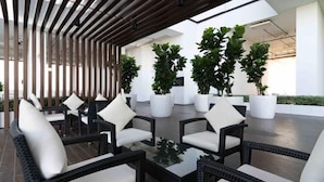 A modern outdoor seating area with wicker chairs and white cushions, surrounded by potted plants and a wooden slatted ceiling.