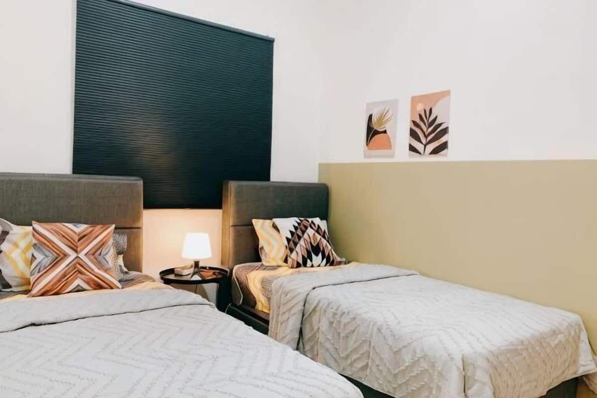 Quarto de hotel com duas camas de solteiro, uma mesa de cabeceira com abajur e uma janela grande com persianas.